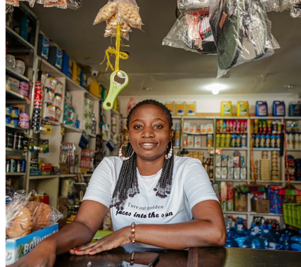 A shop owner smiling behind her counter, with shelves of goods behind her